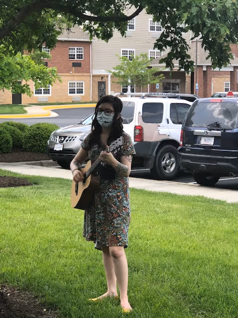 A woman wearing a face mask and a floral dress is standing barefoot on grass playing an acoustic guitar outdoors. Behind her are parked cars and a multi-story residential building with brick and beige siding. Trees and bushes frame the scene.