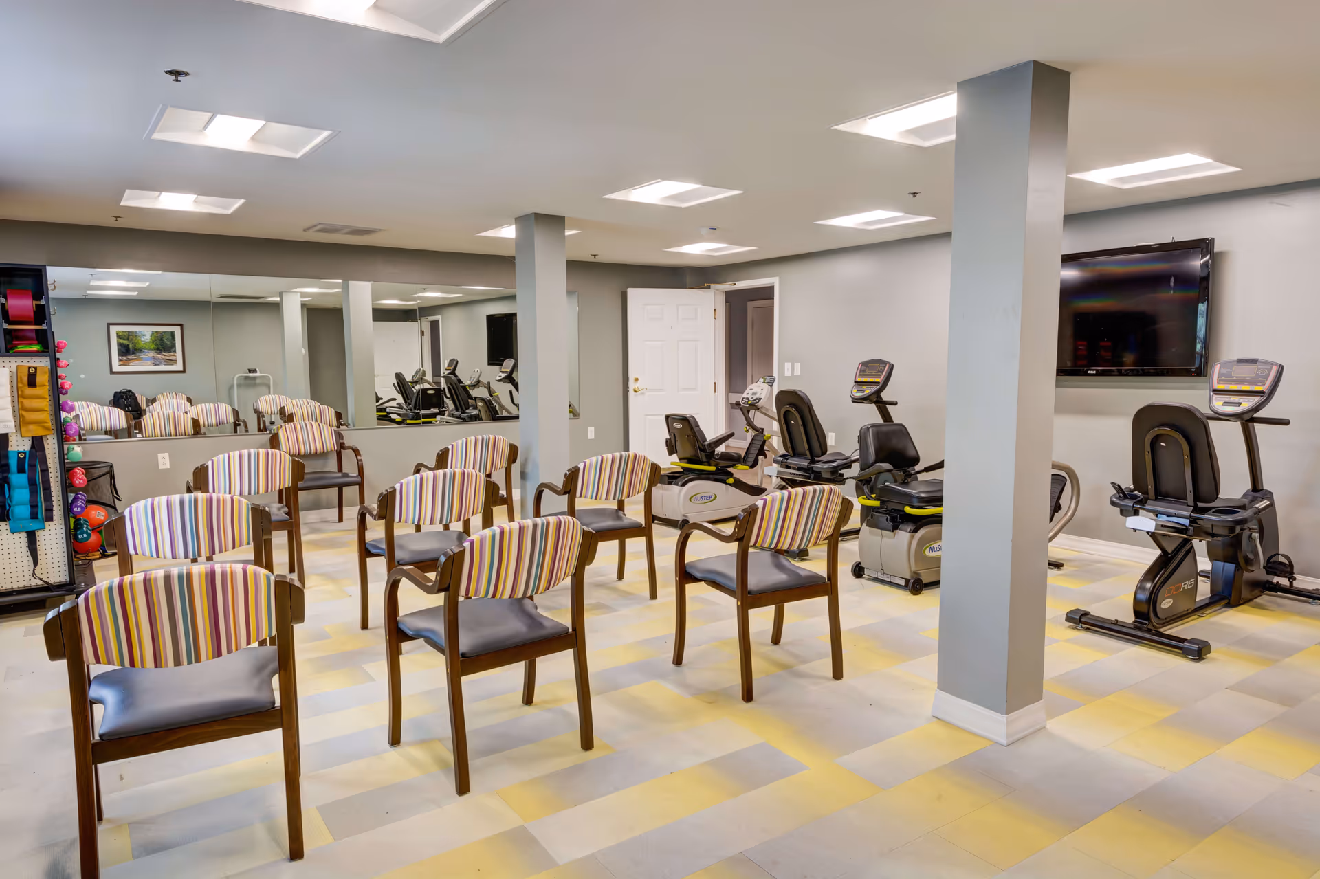 A fitness room with several striped cushioned chairs arranged in rows facing exercise equipment including recumbent bikes and a wall-mounted TV. The room has a light gray and yellow checkered floor, gray walls, and a large mirror along one side. Exercise bands and weights are visible on a rack to the left.