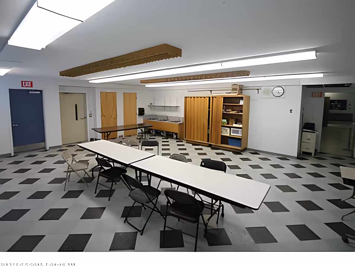 A large room with a checkered black and white floor, several folding tables arranged in a row with folding chairs around them. The room has white walls, fluorescent ceiling lights, a clock on the wall, and wooden cabinets with sliding doors. There are two doors on the left side and an open doorway on the right leading to another area.
