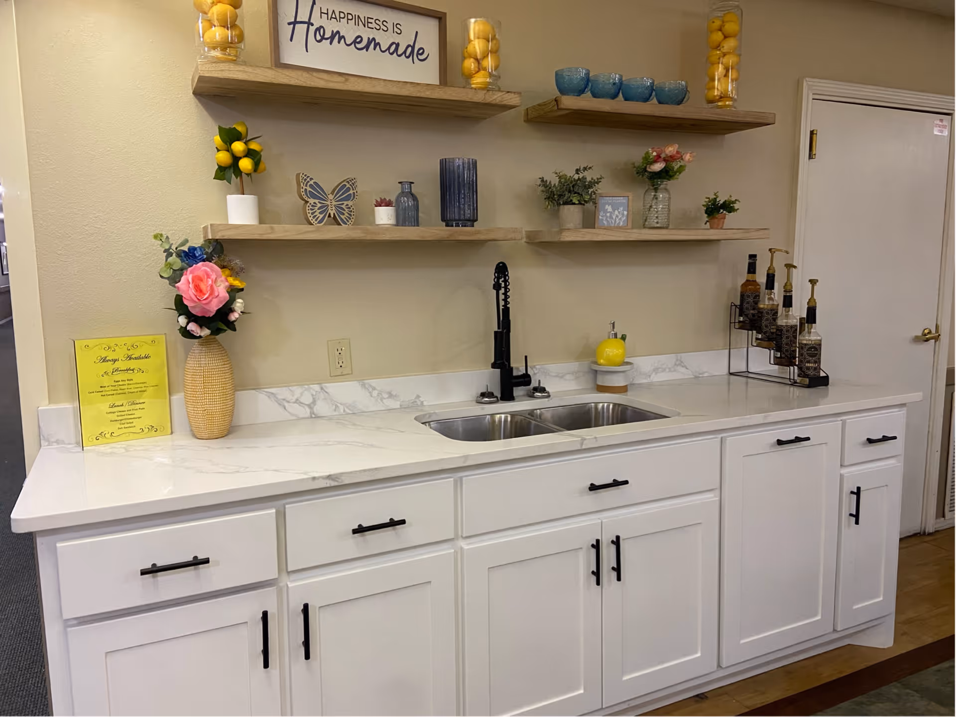 Bright kitchenette with white cabinets, a double sink and decorative open shelves reading 'Happiness is Homemade'.