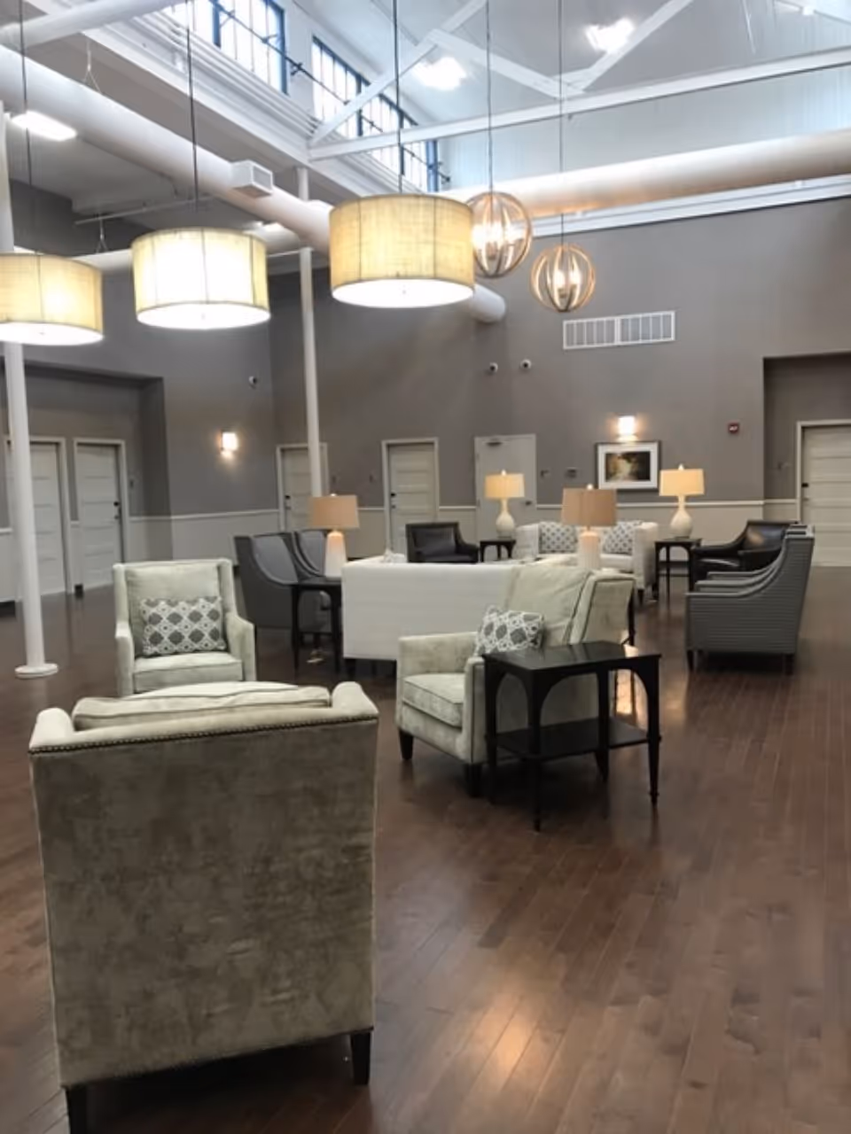 A spacious, well-lit common area with high ceilings and large hanging light fixtures. The room features multiple comfortable armchairs and sofas arranged in small groupings, with side tables and lamps. The walls are painted gray with white trim, and there are several doors along the walls. The floor is wooden, and the overall atmosphere is calm and inviting.