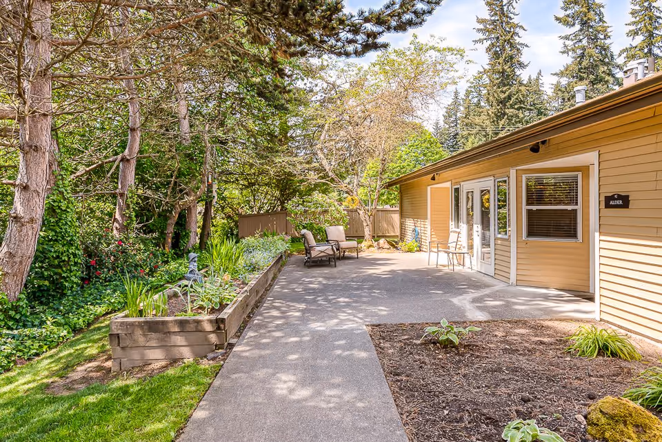 Paved walkway and patio with chairs beside a single-story beige building and landscaped garden under trees.