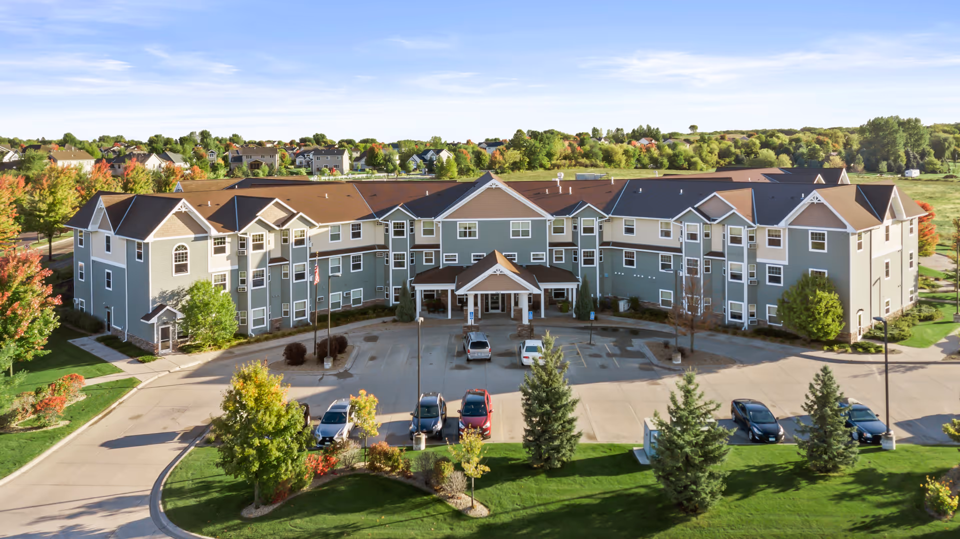 Three-story senior living building with a central covered entrance, parking lot, and landscaped grounds under a blue sky.