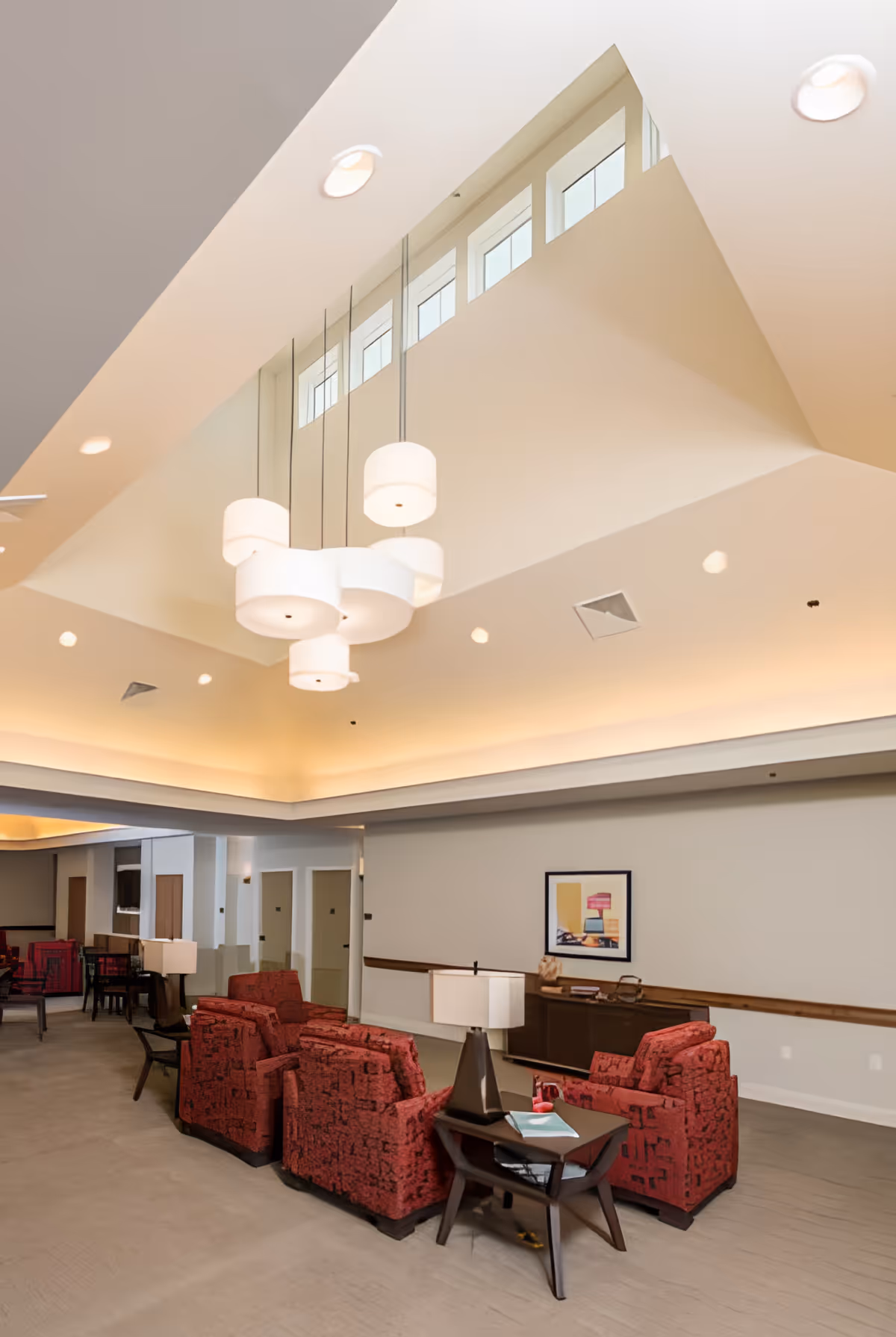 A spacious and well-lit living room area in a senior living facility with high ceilings and modern hanging light fixtures. The room features several red upholstered armchairs arranged around dark wood side tables with lamps. There is a framed artwork on the wall and a wooden console table beneath it. The space has a neutral color palette with beige walls and carpeted floors.
