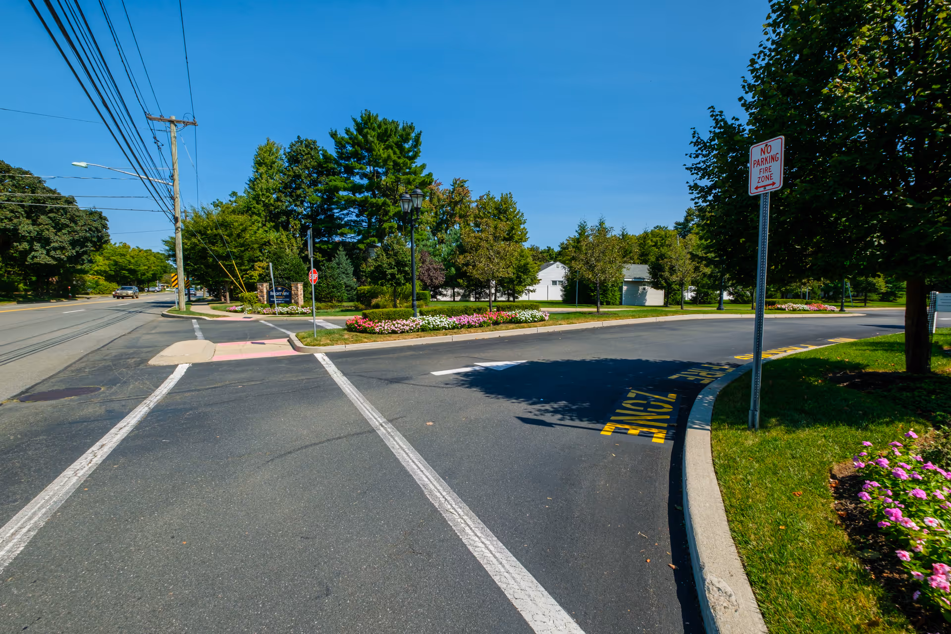 A paved road with a curved driveway entrance surrounded by green grass, trees, and flower beds under a clear blue sky. There is a 'No Parking Fire Zone' sign on the right side of the driveway and utility poles with power lines on the left side of the road.
