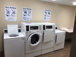 Laundry room with four white laundry machines including a top-loading washing machine, front-loading washing machine, and two dryers. Three instructional posters about washing and drying clothes are displayed on the beige wall above the machines. A folding table is visible on the right side of the room.