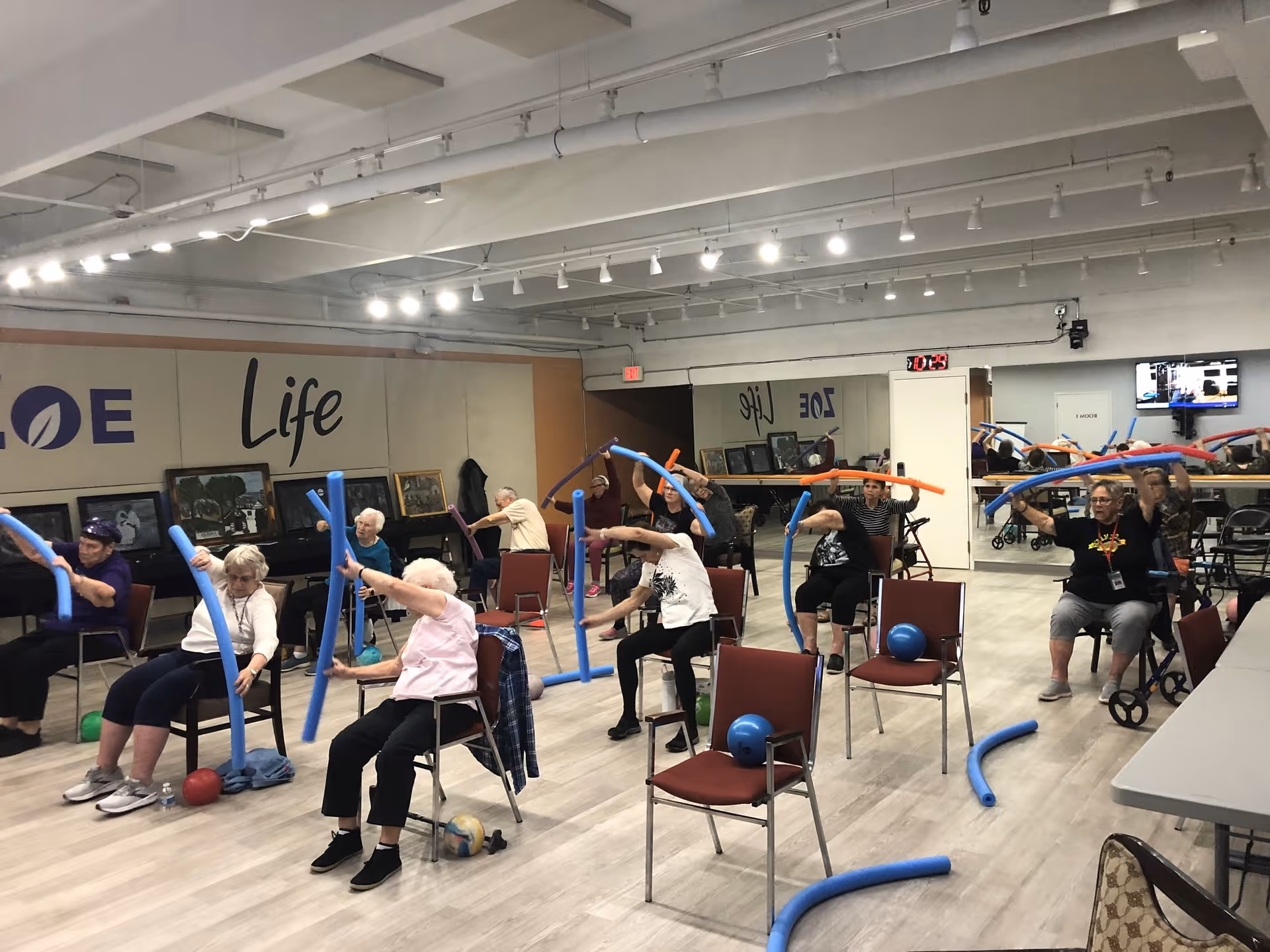 A group of elderly individuals seated on chairs in a spacious room participating in a seated exercise class using colorful foam noodles. The room has light-colored flooring, a mirrored wall, and framed pictures along one side. The words 'Life' and 'ZONE' are visible on the wall.