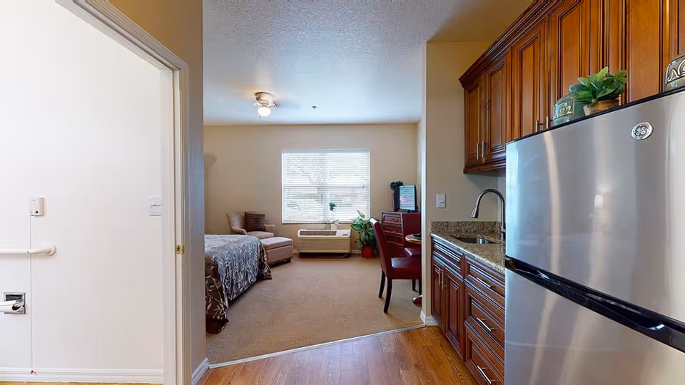 View of a senior living facility room at Chapman Manor at Lutz showing a small kitchenette with wooden cabinets and a stainless steel refrigerator on the right. The room extends into a living and sleeping area with a bed, an armchair, a window with blinds, an air conditioning unit below the window, a small table with two red chairs, and a TV on a wooden stand. The left side shows a partial view of a bathroom door with grab bars and a toilet paper holder.
