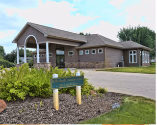 Exterior view of a single-story building with a covered entrance supported by white columns. The building has a brown exterior with stone accents and multiple windows. In front of the building, there is a landscaped area with green shrubs and a sign that reads 'Rollins Head Community Center 1438 Cascade Drive'. The sky is partly cloudy and there are trees in the background.