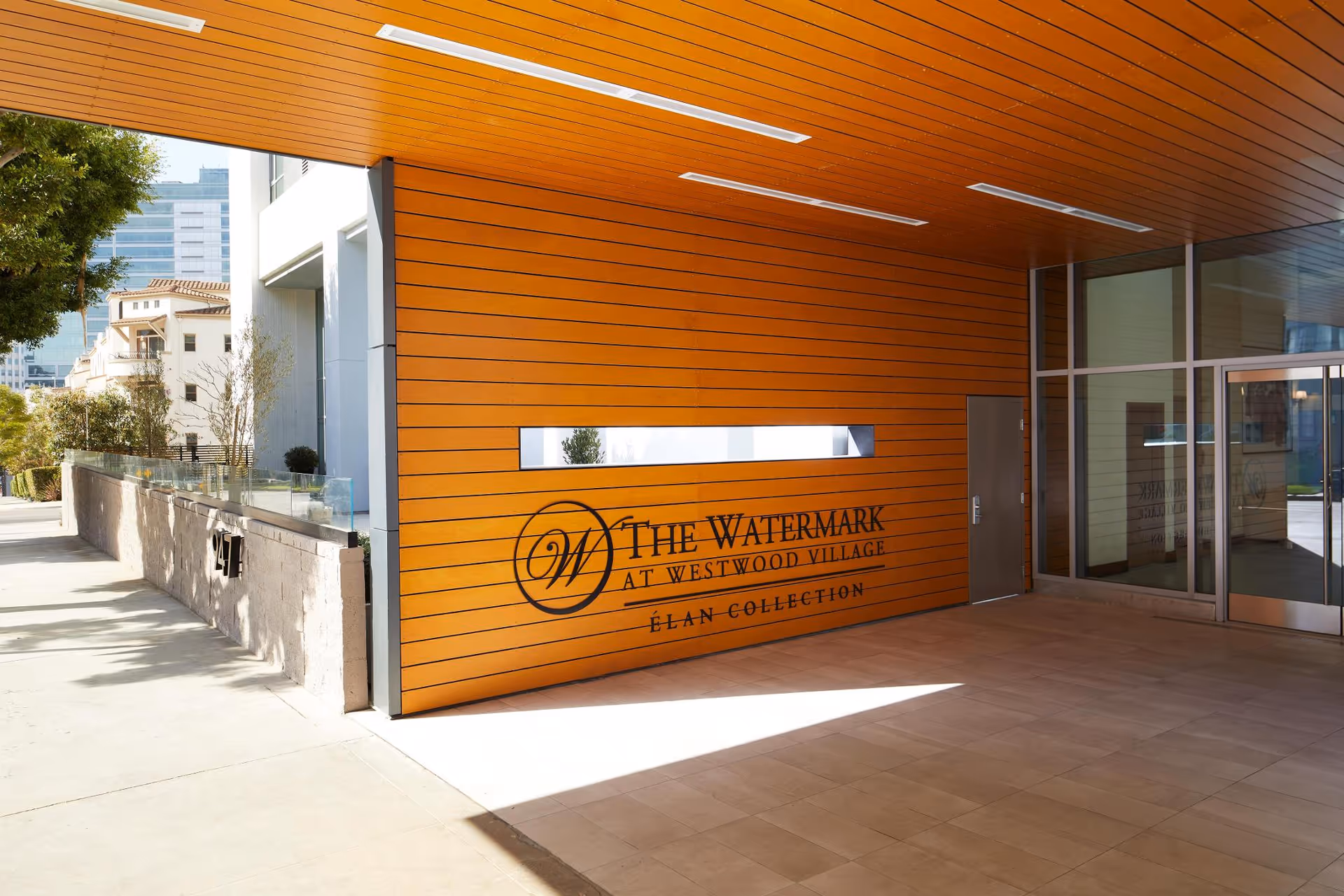 Entrance area of The Watermark at Westwood Village featuring a modern wooden panel wall with the facility's name and logo, a glass door entrance, and a sidewalk with trees and buildings in the background.