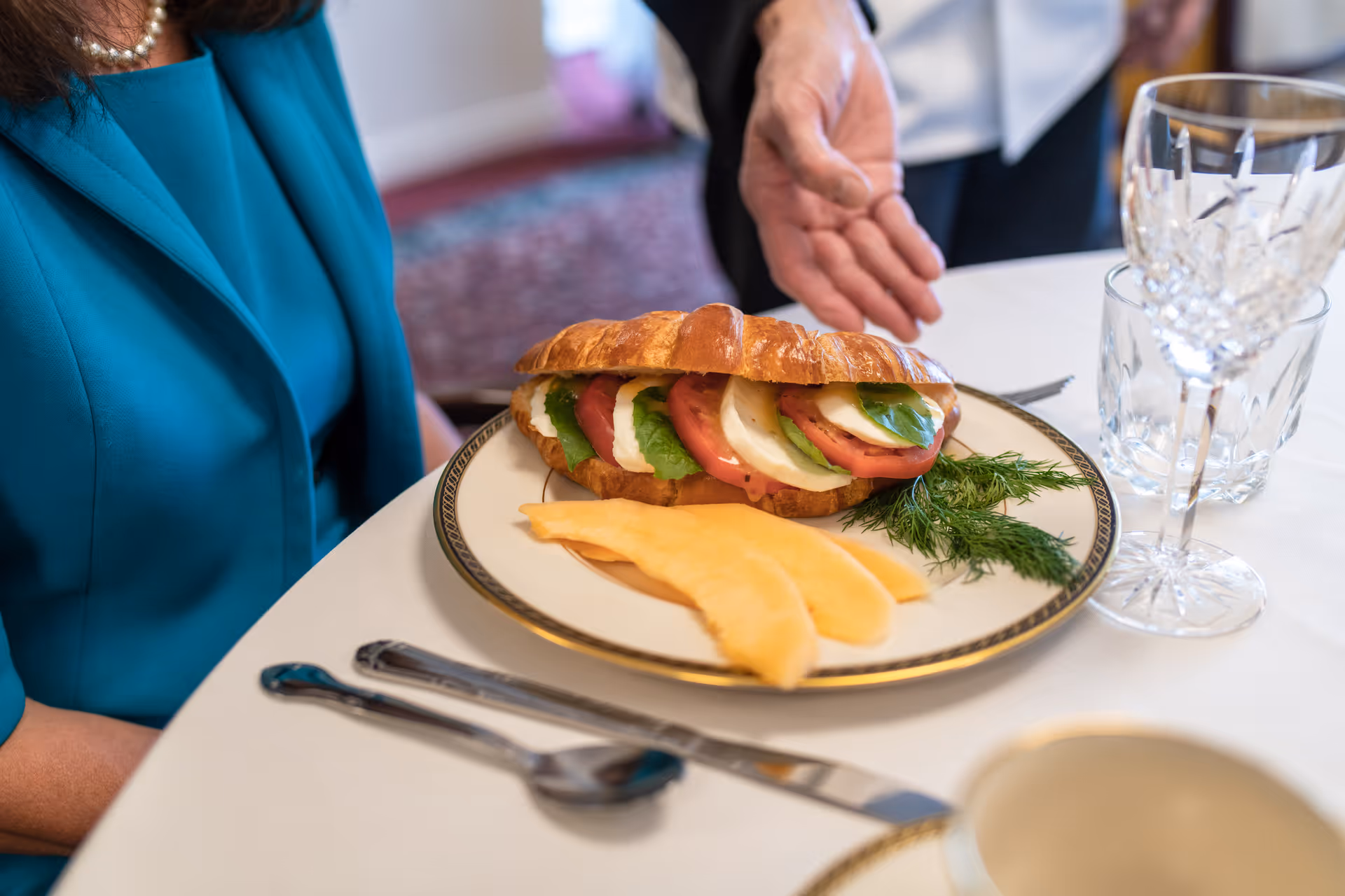 A close-up of a plate with a croissant sandwich filled with tomato, mozzarella, and basil, accompanied by slices of cantaloupe and a sprig of dill. A person in a teal outfit is seated at the table, and another person's hand is gesturing towards the plate. The table is set with crystal glassware and silverware on a white tablecloth.