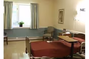 A senior living bedroom with an adjustable bed, overbed table, armchair, window with curtains, and framed wall art.