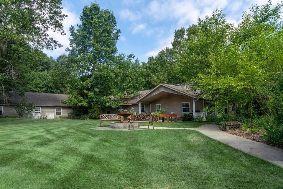 A peaceful outdoor garden area at Golden Orchards featuring a well-maintained green lawn, a central multi-tiered water fountain, benches around the fountain, and a single-story building surrounded by trees under a partly cloudy sky.