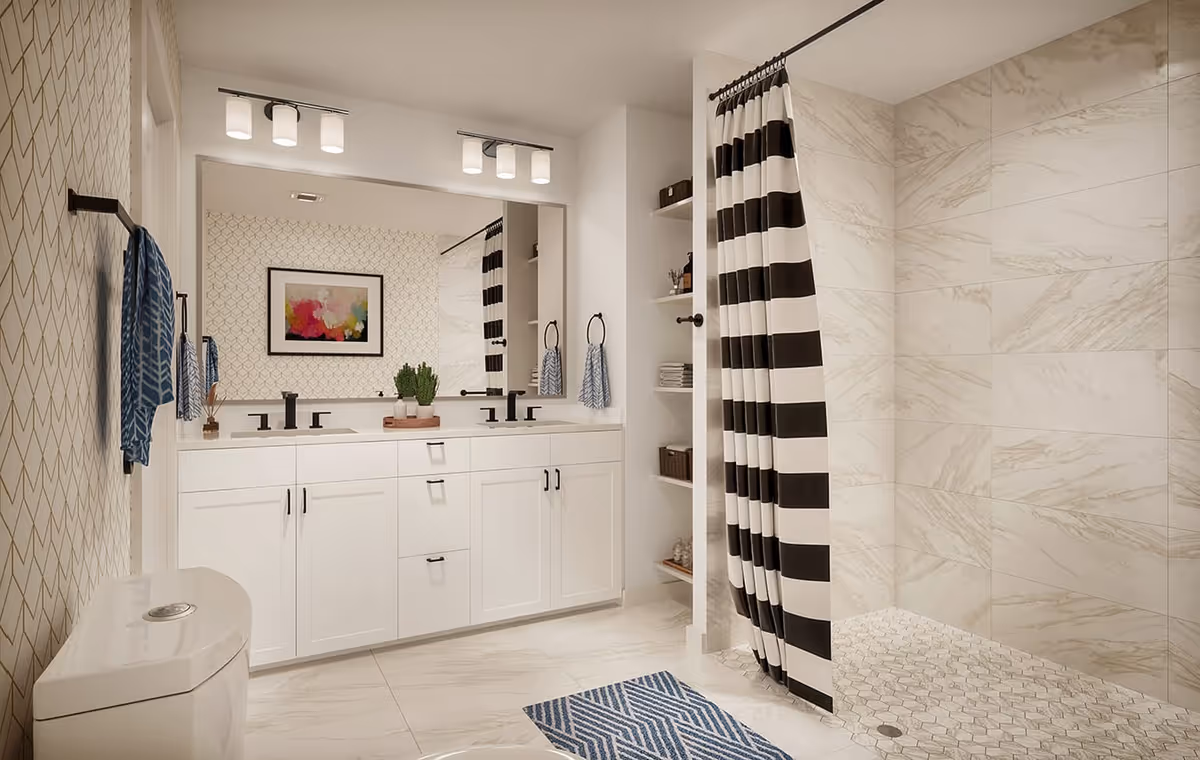 Modern bathroom with white cabinetry and double sinks, large mirror with two light fixtures above, a toilet, a shower area with black and white striped curtain, beige marble tiles on walls and floor, blue patterned rugs, and decorative items including a framed abstract painting and plants.