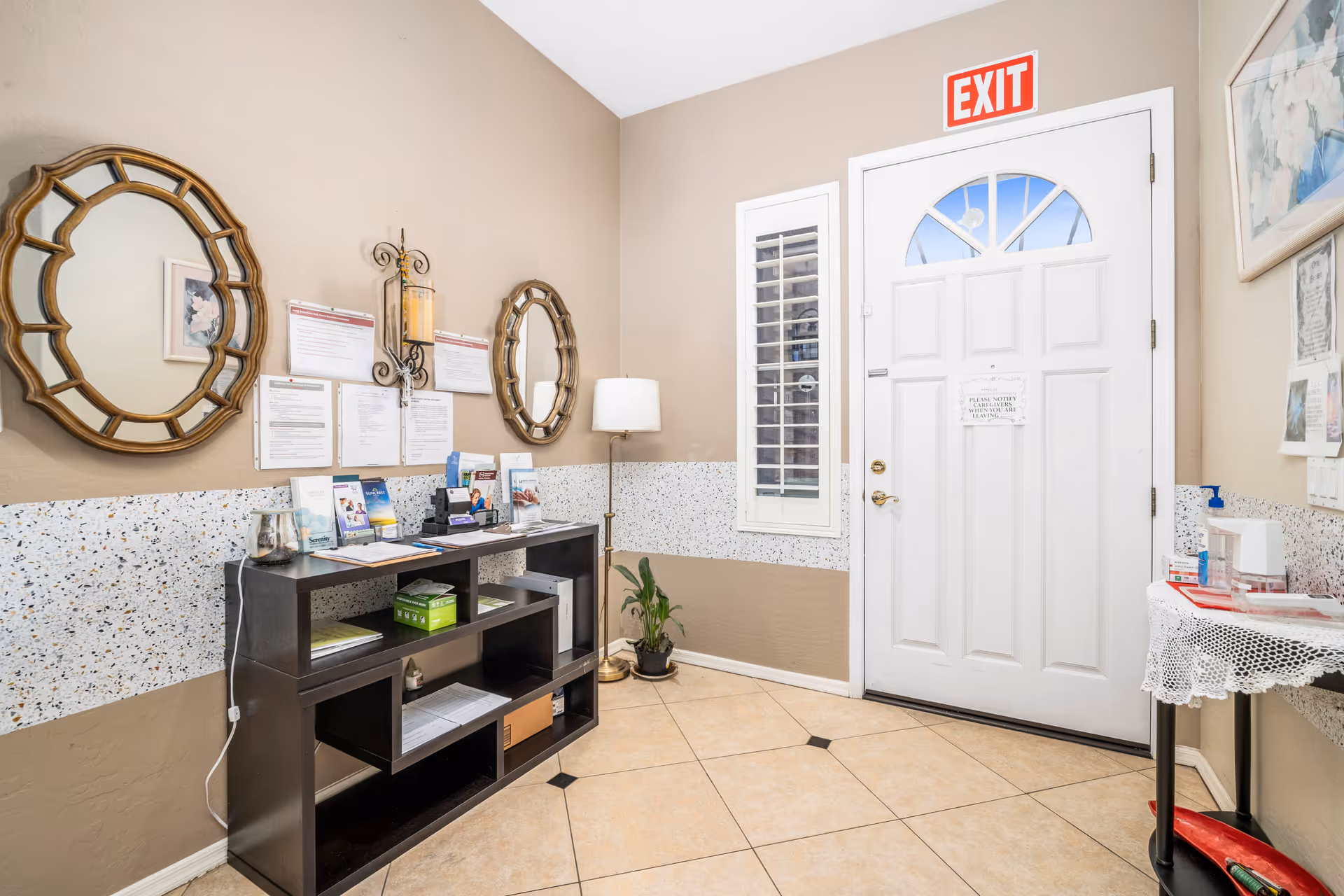Interior view of an entryway or lobby area in an assisted living facility. The space features a white door with an exit sign above it, two decorative round mirrors on the left wall, a dark wooden shelf holding brochures and documents, a floor lamp, a small potted plant, and a small table with a lace cover holding hand sanitizer and other items.