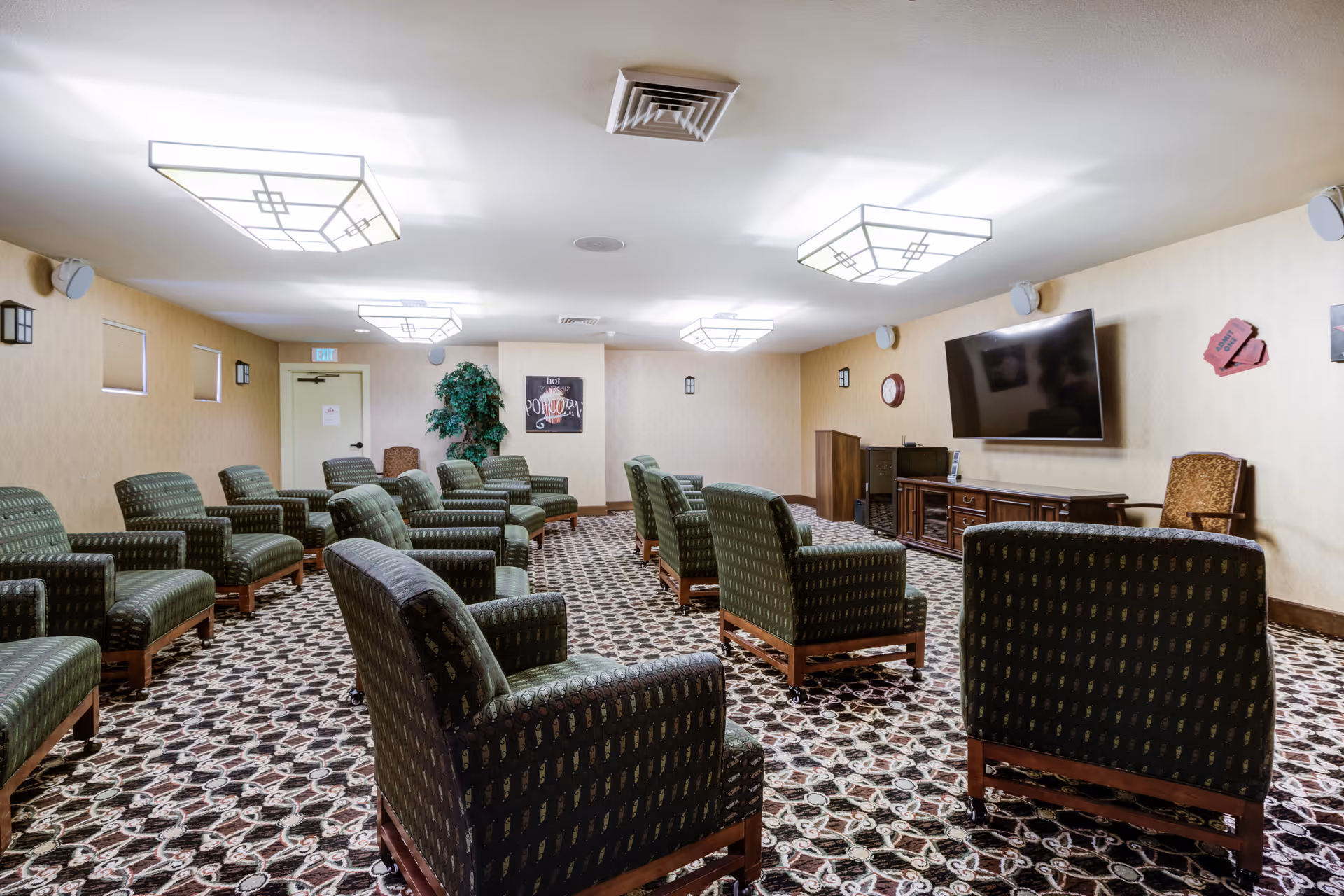 A senior living community lounge with rows of green upholstered armchairs arranged facing a wall-mounted TV and media cabinet.