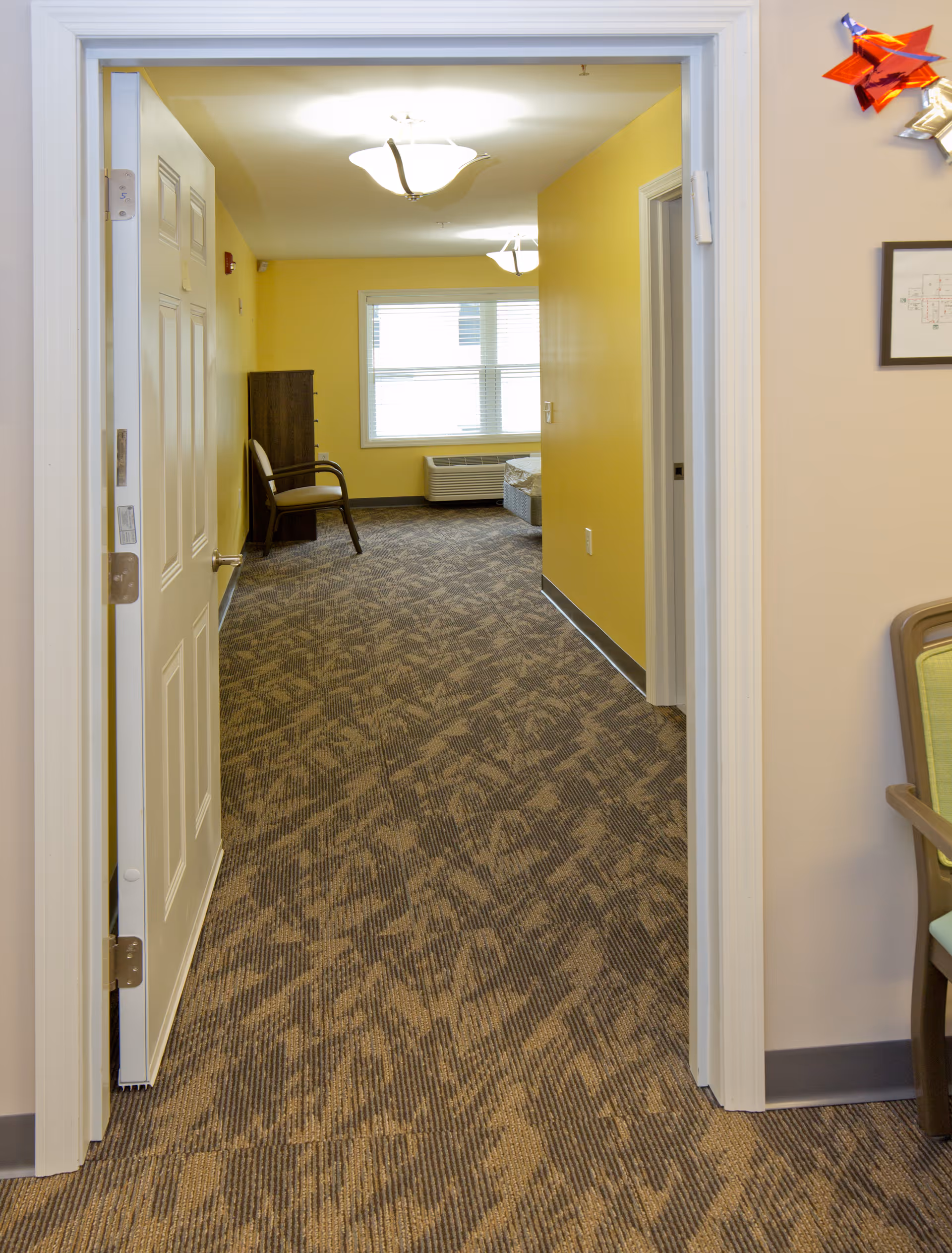 View through an open doorway into a room with yellow walls, patterned carpet, a window with blinds, a wooden chair, and a dresser. The room is well-lit with ceiling lights.