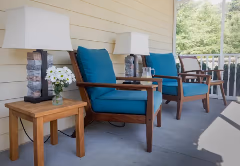 A porch area with two wooden chairs featuring blue cushions, a small wooden side table with a vase of white daisies, two stone-based table lamps, and a pitcher with a glass of lemonade. The porch has a railing and overlooks greenery.