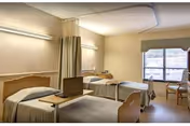 A hospital-style room with two single beds separated by a curtain. Each bed has a wooden headboard and is covered with beige bedding. A small table with a laptop is placed on the bed in the foreground. The room has beige walls, two fluorescent lights above the beds, a window with a beige valance, and two chairs near the window.