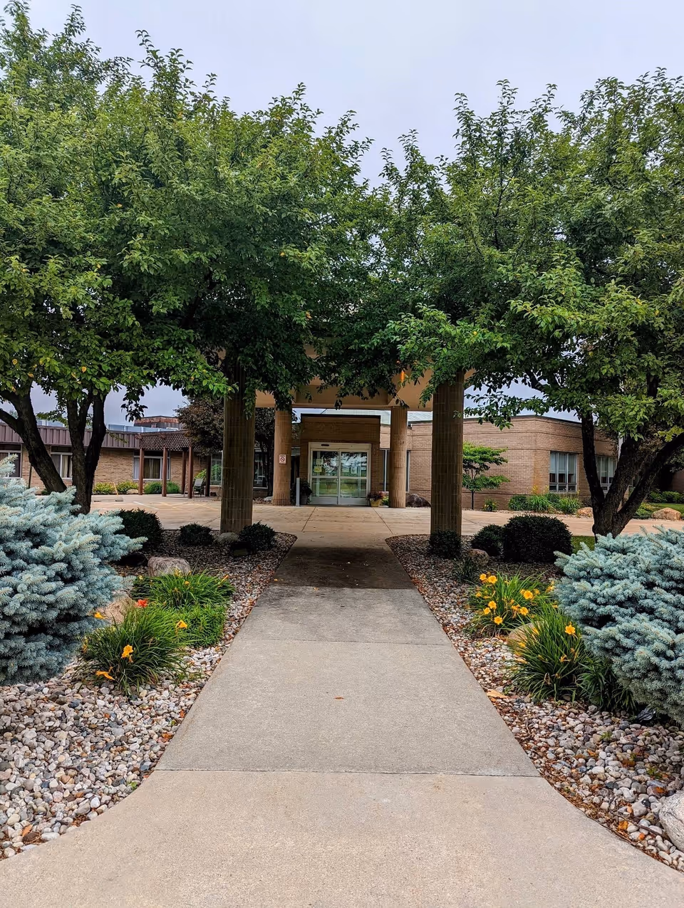 A concrete walkway leads through a landscaped garden with green trees, shrubs, and yellow flowers towards the entrance of a single-story brick building with glass doors under a covered entryway.