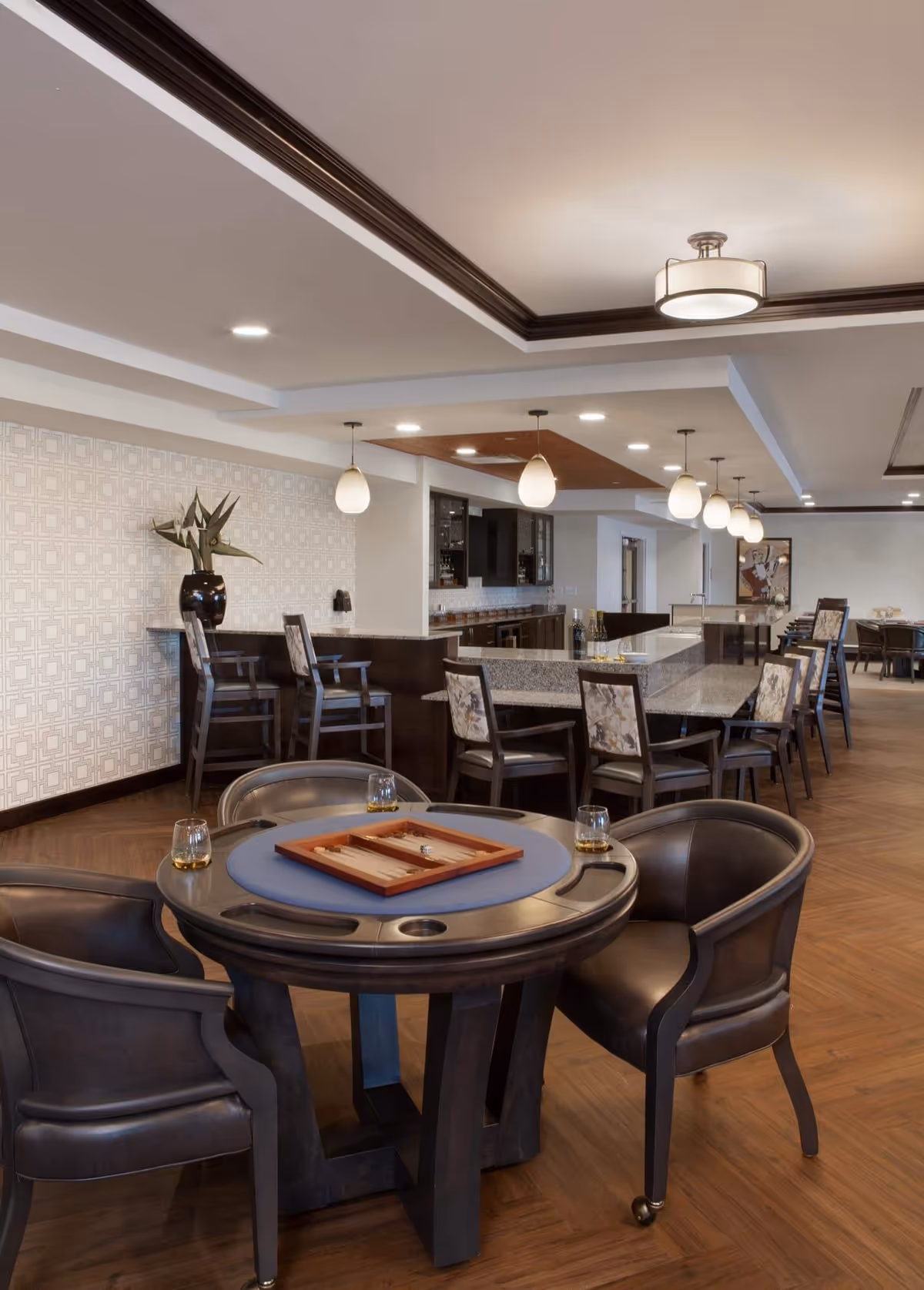A spacious and well-lit common area with a round game table featuring a backgammon set and four leather chairs. In the background, there is a long granite countertop with high chairs, pendant lights hanging from the ceiling, and a decorative vase with plants on a side counter. The room has wooden flooring and a modern ceiling design with recessed lighting.