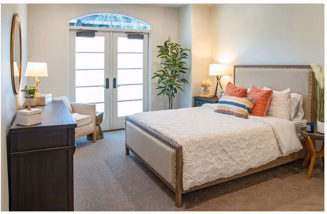 A bright and cozy bedroom featuring a large bed with a beige upholstered headboard, white textured bedding, and decorative pillows in orange and striped patterns. The room includes a dark wooden dresser with a round mirror above it, a beige armchair, two bedside tables with lamps, a tall green plant in the corner, and double doors with frosted glass panels letting in natural light.