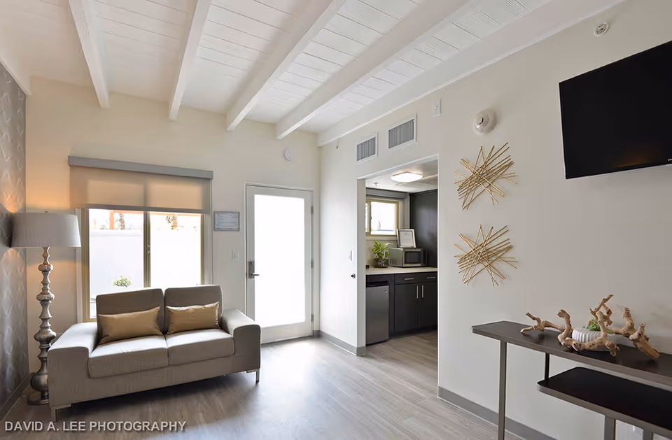 Bright living room with a sofa, floor lamp, wall-mounted TV, decorative wall art and a view into a small kitchenette.