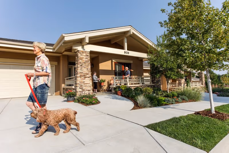 An elderly woman walking a small brown dog on a red leash on a sidewalk in front of a single-story residential building with a porch. Two other elderly people are standing and sitting on the porch, which has stone pillars and potted plants. The building number 9170 is visible above the porch. The scene is sunny with a clear blue sky and green landscaping including trees and grass.