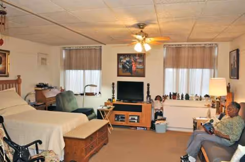 Senior living bedroom with a bed, TV, seating, and a resident reading.