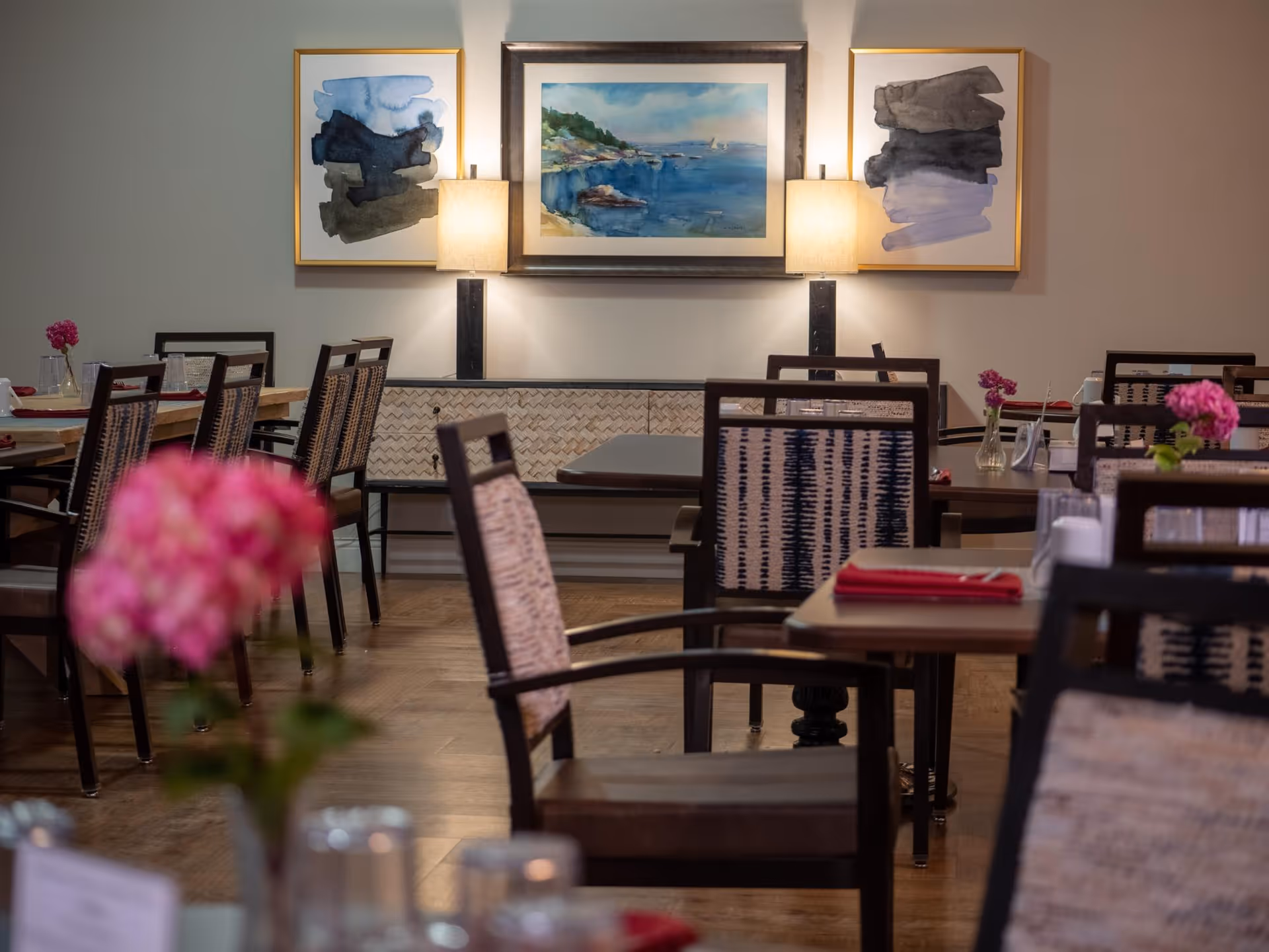 Neatly arranged dining room with wooden tables and patterned chairs, wall art and lamps, and small floral centerpieces on the tables.
