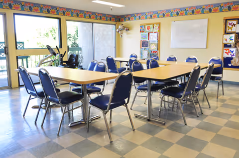 A bright room with two rectangular tables pushed together surrounded by blue cushioned chairs. The room has large windows letting in natural light, colorful artwork on the walls, and a patterned border near the ceiling. The floor has a checkered tile pattern.
