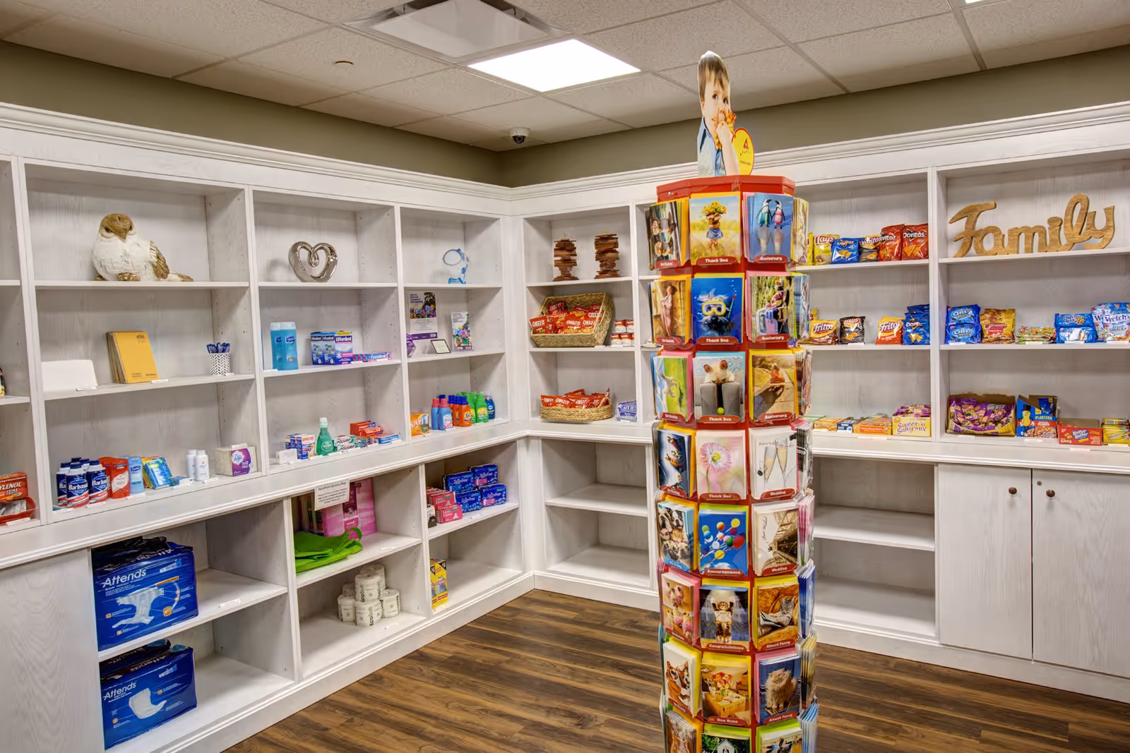 Interior view of a small store or shop area with white shelves stocked with various personal care products, snacks, and other items. A rotating display rack in the center holds greeting cards featuring animals and other images. The word 'Family' is displayed on one of the shelves.