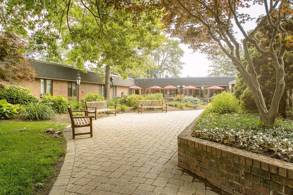 Paved courtyard with wooden benches, planted landscaping, and a low brick building with red patio umbrellas in the background.