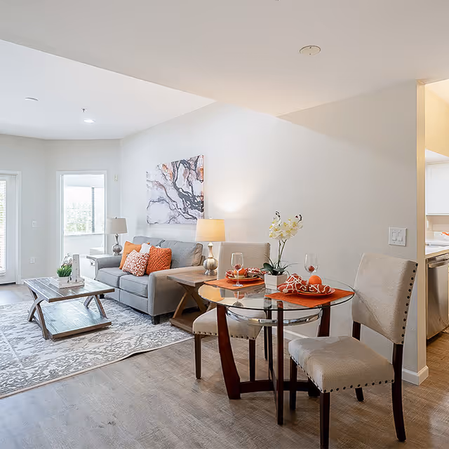 A bright and modern living room and dining area with a gray sofa adorned with orange and patterned pillows, a wooden coffee table on a patterned rug, a side table with a lamp, and a round glass dining table set with two beige upholstered chairs, orange placemats, wine glasses, and a small potted orchid. The room has light-colored walls and wood flooring, with natural light coming through windows in the background.