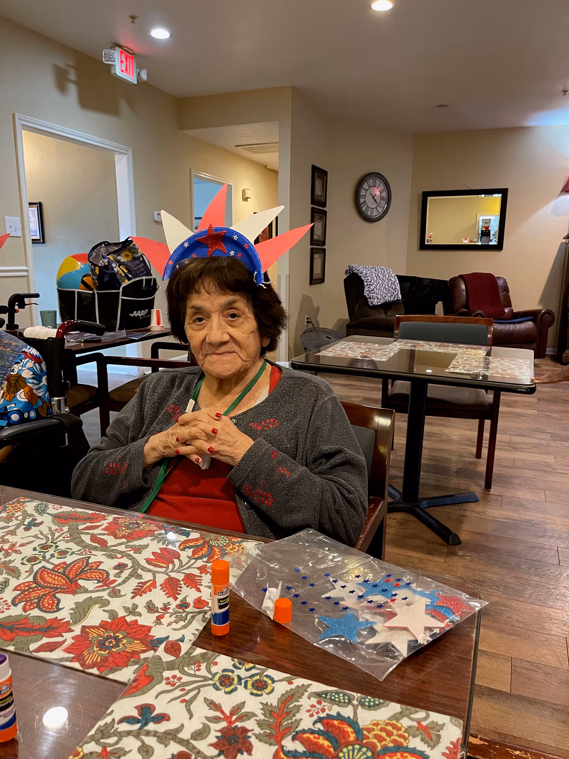 An elderly woman sitting at a table in a common area, wearing a handmade red, white, and blue paper crown with stars. The table has floral placemats and crafting supplies including glue sticks and star-shaped decorations. In the background, there are chairs, a clock on the wall, and a mirror reflecting part of the room.
