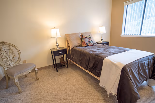 A simple bedroom with a single bed covered in a brown blanket and a white throw at the foot. There are two bedside tables with lamps on each side of the bed. A decorative chair is placed against the wall, and a window with vertical blinds lets in natural light.