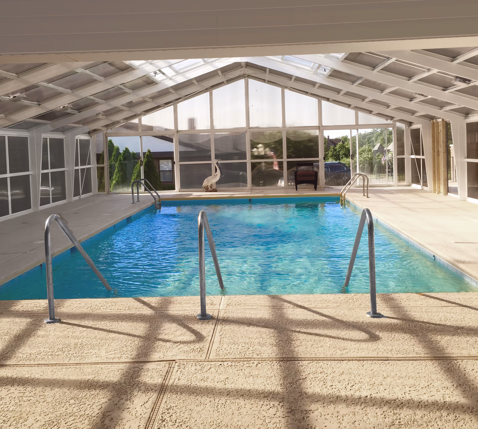 An indoor enclosed swimming pool with metal handrails, skylit vaulted ceiling, and windows overlooking greenery.
