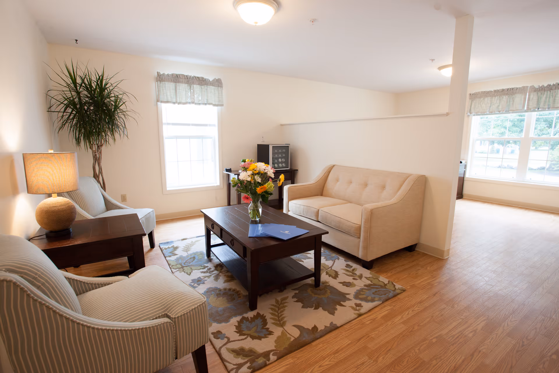 A bright and cozy living room with a beige sofa, two striped armchairs, a wooden coffee table with a vase of colorful flowers, a side table with a lamp, a potted plant, and large windows with light curtains letting in natural light.