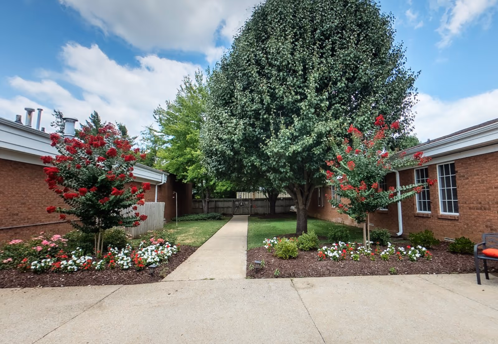 Outdoor courtyard area with a concrete walkway leading through a garden with red and white flowers, two small trees with red blossoms, and a large leafy tree in the center. The area is surrounded by brick buildings under a partly cloudy blue sky.