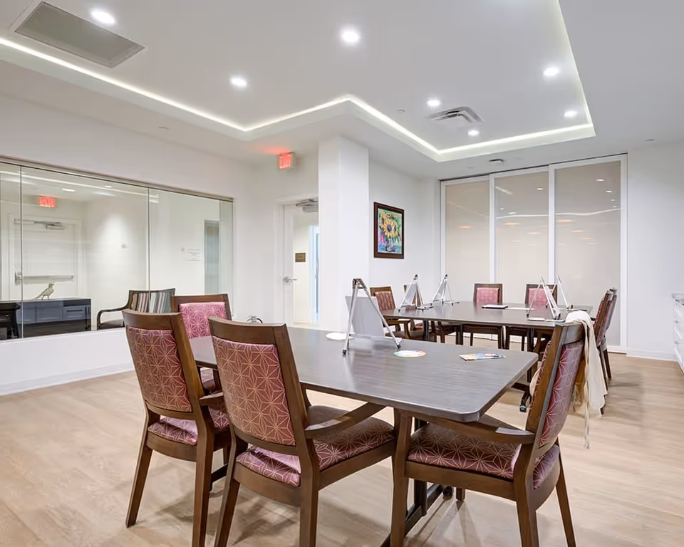 A bright and modern common area with two rectangular tables surrounded by wooden chairs with patterned pink cushions. The room has light wood flooring, white walls, and a ceiling with recessed lighting and a decorative light strip. There is a colorful framed painting on one wall and large glass windows and doors in the background.