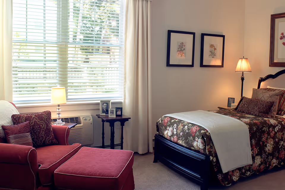 A cozy bedroom with a floral-patterned bedspread on a black wooden bed frame, a red armchair with matching ottoman, a small side table with framed photos, and two framed floral prints on the wall. The room is softly lit by a table lamp and a floor lamp, with a large window covered by white blinds and curtains.