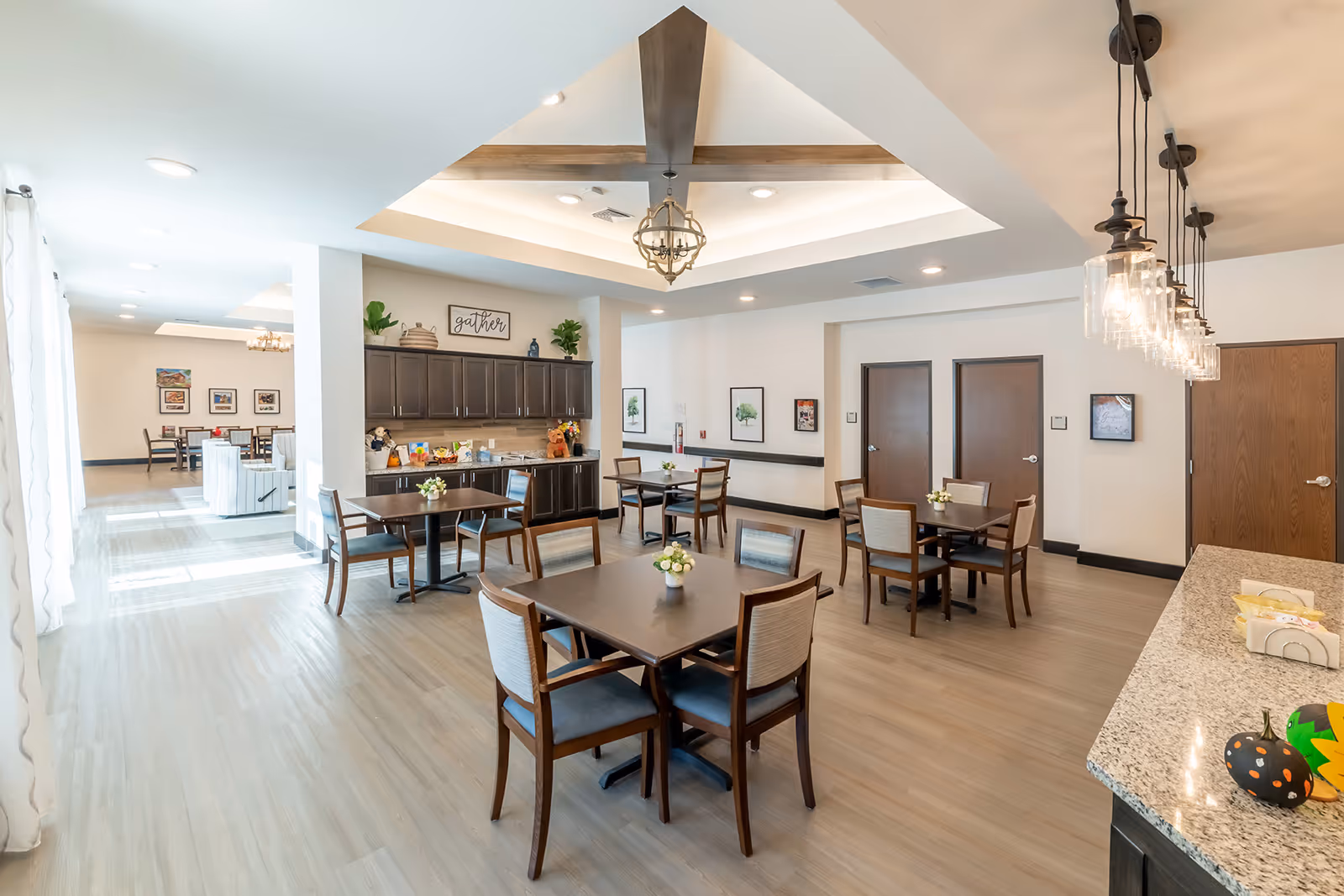 Bright communal dining area with multiple tables and chairs, cabinetry and a serving counter with pendant lights.