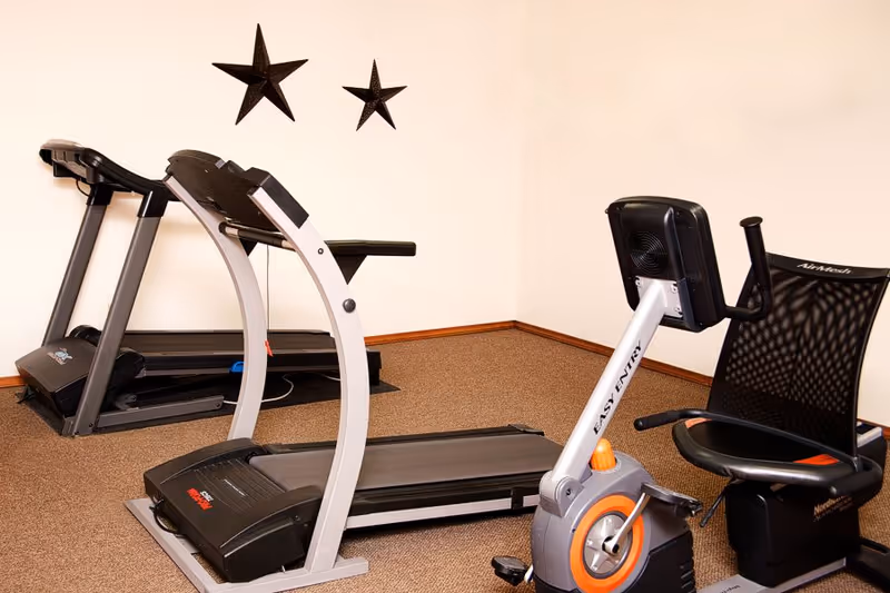 A small exercise room with two treadmills and a stationary recumbent bike. The room has beige walls with two decorative star-shaped wall hangings and brown carpeted flooring.