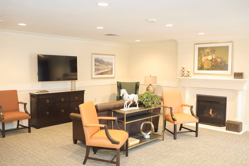 A cozy senior living common area featuring a fireplace with a floral painting above it, a flat-screen TV mounted on the wall, a dark wooden cabinet, and several chairs including two orange armchairs and a green wingback chair. A small table with decorative items such as a white horse figurine and a plant is placed in the center.