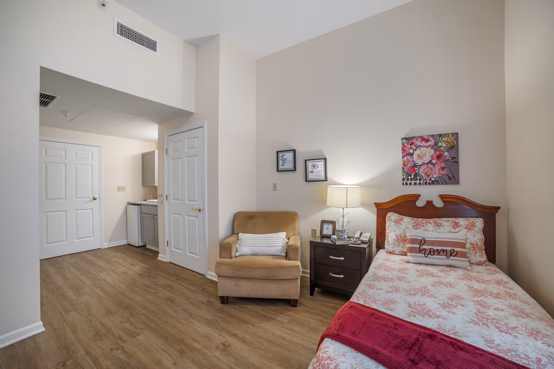 A bright senior-living bedroom with a single bed, bedside table and lamp, an armchair, and a small kitchenette visible in the alcove.