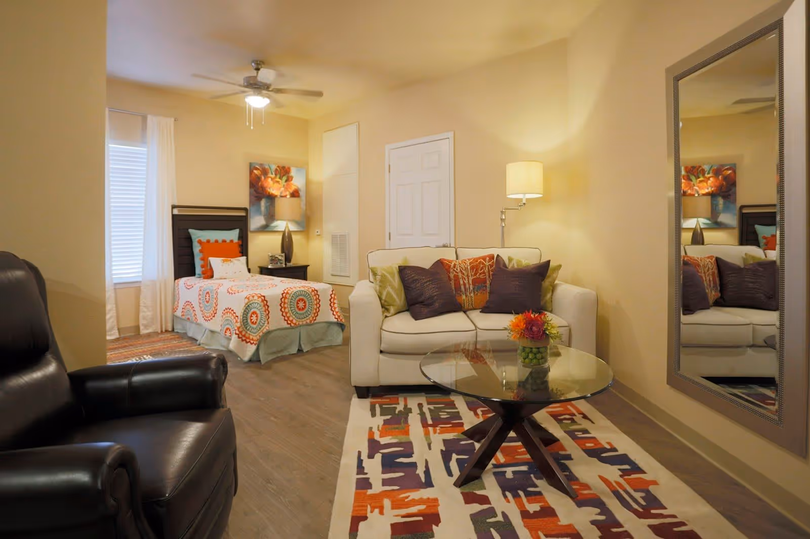 A cozy living and bedroom area in a senior living facility with a white sofa adorned with colorful pillows, a glass coffee table with a small flower arrangement, a large mirror on the wall, a black leather armchair, and a single bed with a patterned bedspread and pillows. The room has warm lighting and a ceiling fan.
