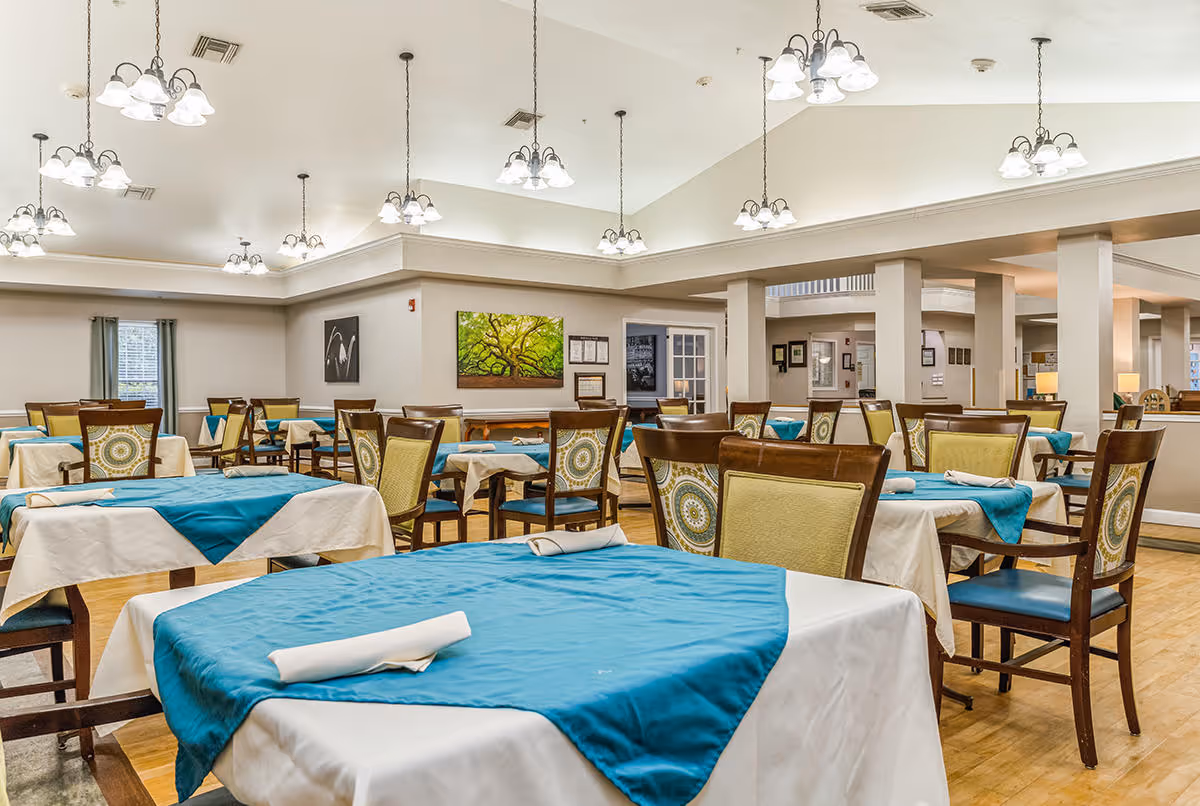 A spacious dining room with multiple tables covered in white tablecloths and teal table runners. Each table is set with a rolled white napkin. The chairs have wooden frames with patterned cushions. The room is well-lit with multiple hanging light fixtures and has light-colored walls and hardwood floors. Artwork and windows are visible on the walls.