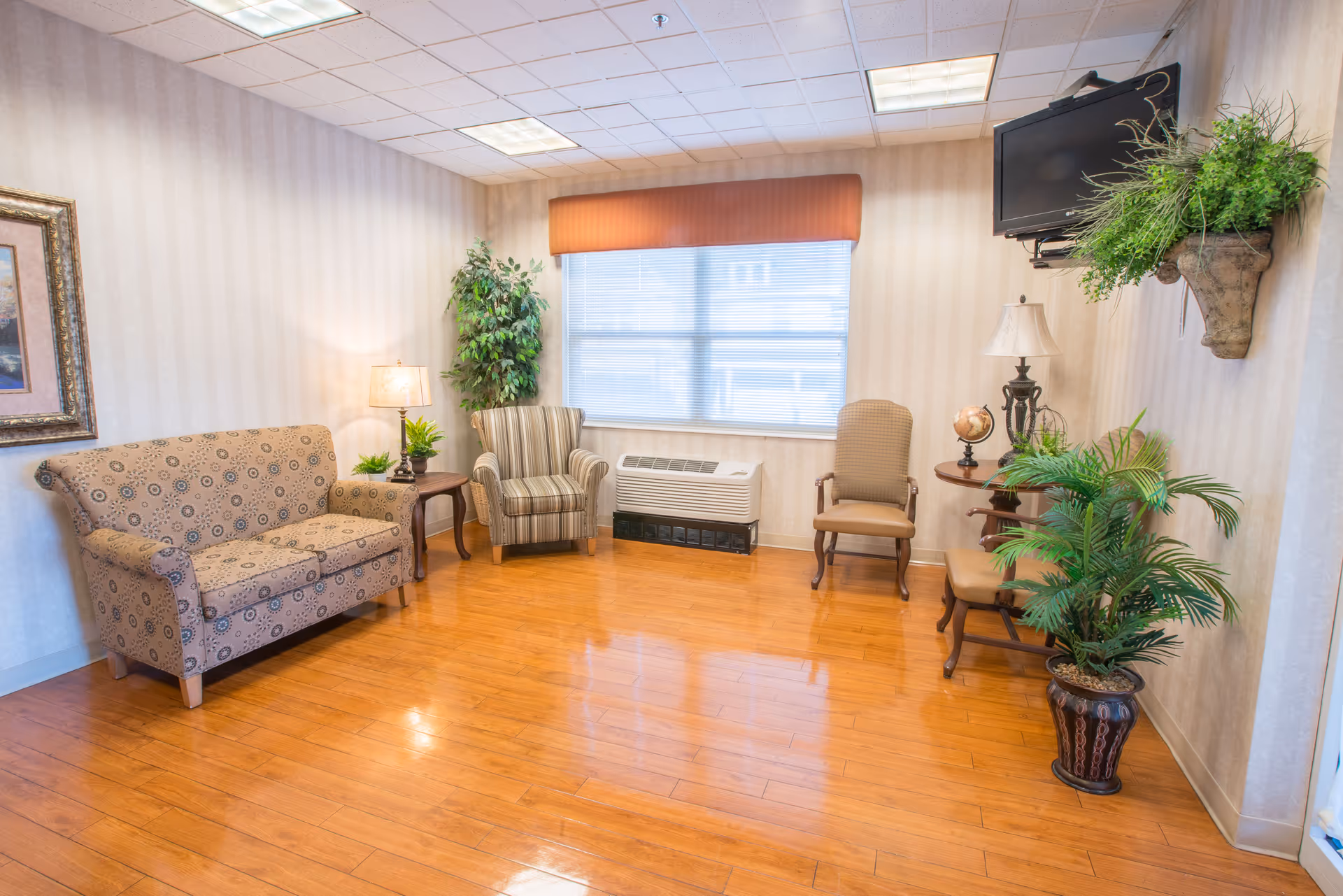 A cozy living room with wooden flooring, a patterned sofa, a striped armchair, two wooden chairs, a small round table with a lamp and a globe, several potted plants, a wall-mounted TV, and a large window with blinds and an orange valance.