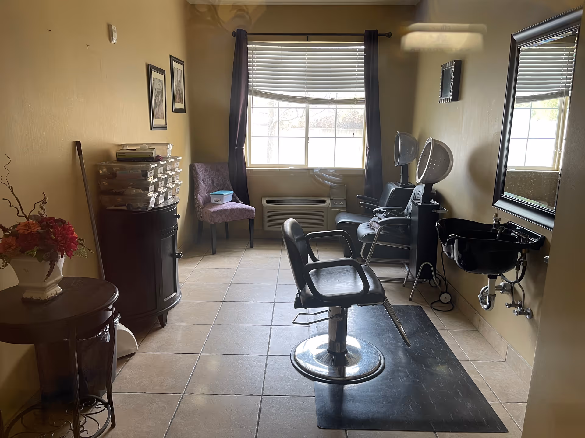 Interior view of a small hair salon room with a black salon chair on a black mat in the center, two black hair dryer chairs with hooded dryers against the right wall, a black sink with a mirror above it, a purple upholstered chair near a window with blinds and dark curtains, a small cabinet with organized plastic drawers, and a small round table with a flower arrangement on the left side.