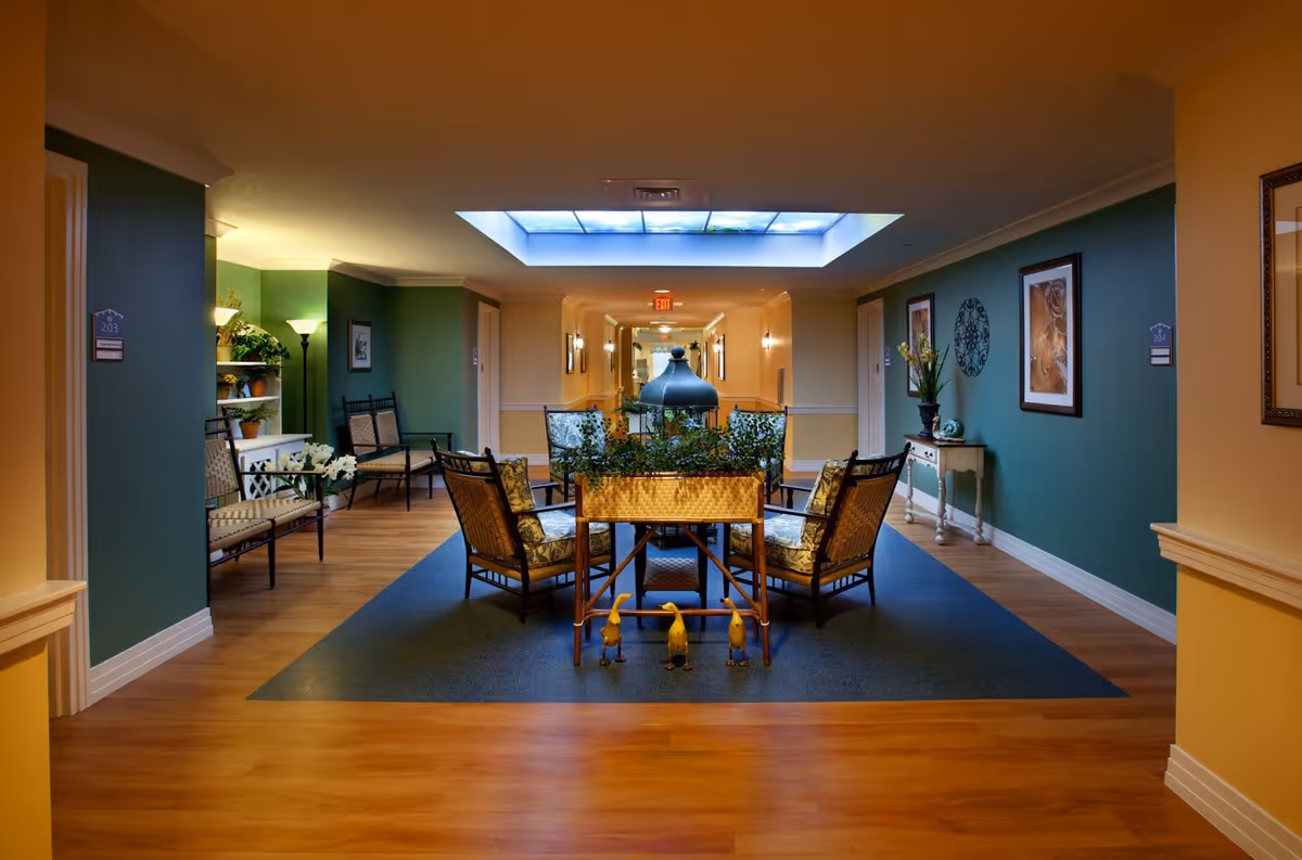 A cozy seating area in a senior living facility hallway with cushioned chairs arranged around a central table decorated with plants. The walls are painted green with framed artwork and decorative items, and there is a skylight above providing natural light. The floor is wood with a carpeted section under the seating area.